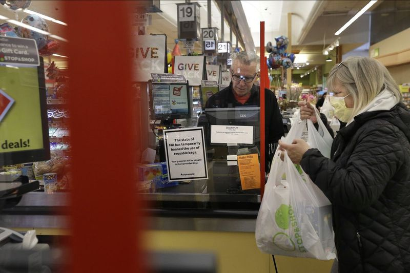 Kassören George Wallace, i mitten, arbetar bakom en plastsköld som en shoppare, till höger, placerar matvaror i en vagn på torsdag i en livsmedelsbutik i Quincy, Mass.