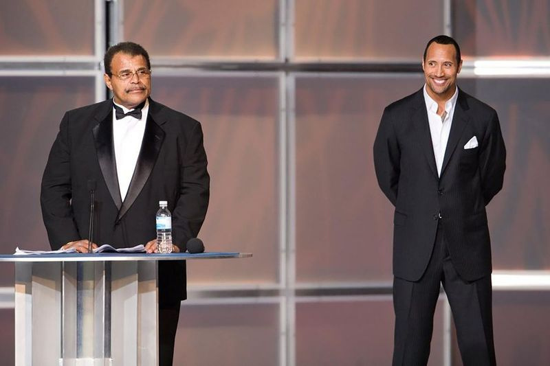 En esta foto de 2008 proporcionada por WWE, Inc., Rocky Soul Man Johnson, a la izquierda, habla en su ceremonia de inducción al Salón de la Fama de la WWE, mientras su hijo, Dwayne The Rock Johnson, observa.