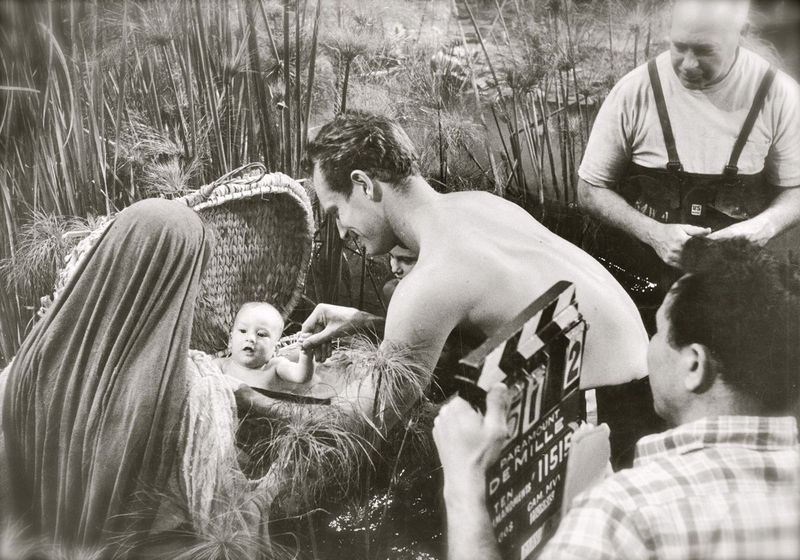 Ako sa pozerá režisér Cecil B. DeMille (v pozadí), o malého Mosesa, ktorého hrá Fraser Heston, sa stará jeho slávny skutočný otec Charlton Heston počas tejto scény, ktorá sa natáčala na pozadí Paramount Studios v roku 1955.