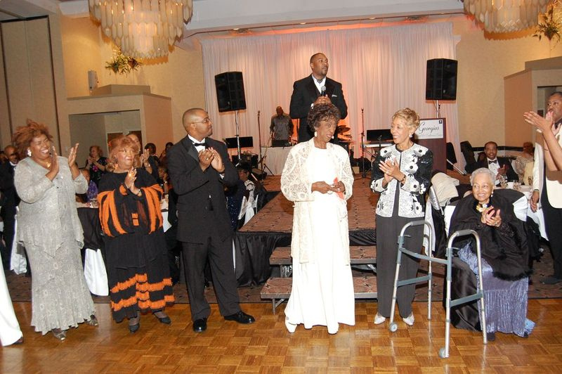 Pr. Henrietta Byrd (din stânga), Pr. Marjorie Cook și Pr. Gaylon McDowell se alătură Pr. Carry, Pr. Evelyn Boyd și Pr. Jeanette Childress la un banchet. Rev. Derrick B. Wells stă pe podium.