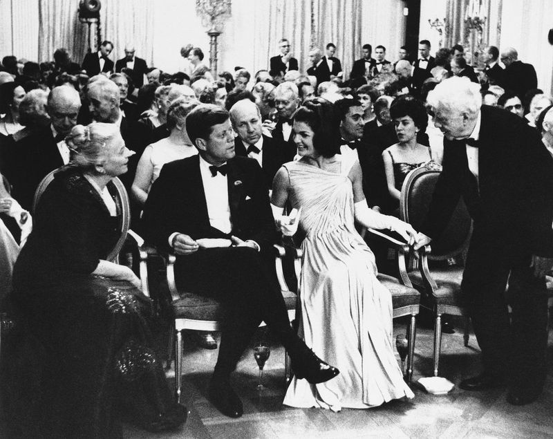 Præsident John F. Kennedy sidder sammen med førstedame Jacqueline Kennedy som digteren Robert Frost, til højre, og Pearl Buck, til venstre, udveksler hilsner i Østrummet i Det Hvide Hus i 1962. Et år tidligere talte Frost ved præsident Kennedys tiltrædelse.
