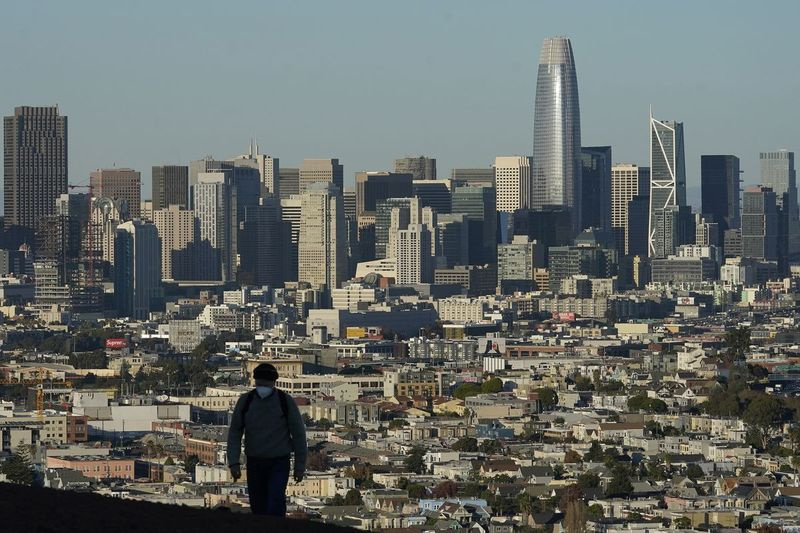 På dette arkivbildet 7. desember 2020 går en person som bærer en beskyttende maske foran skyline på Bernal Heights Hill under koronaviruspandemien i San Francisco.